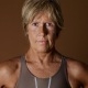 Diana Nyad with short hair wearing a sleeveless top stands against a brown background.